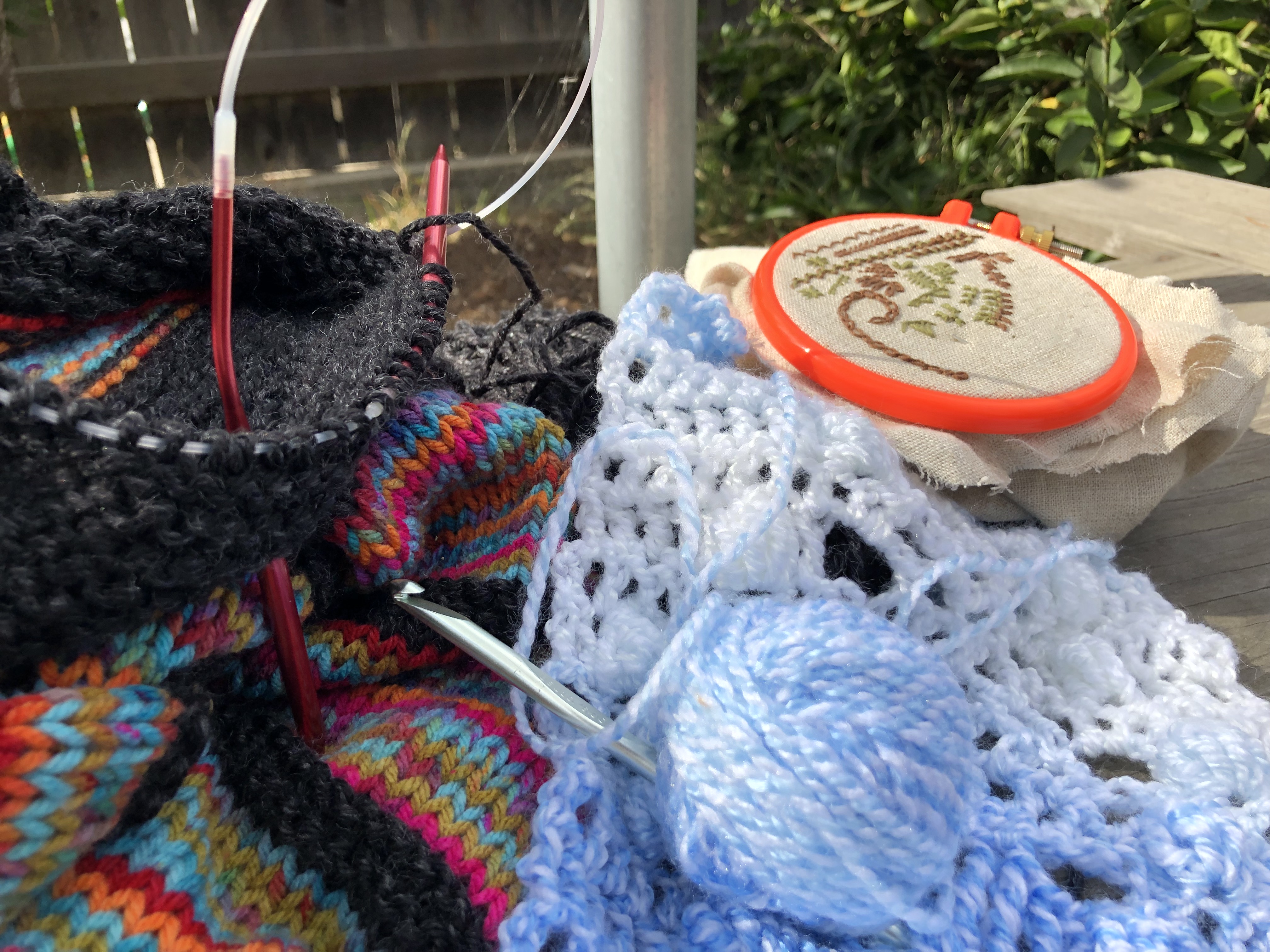 Yarn, knitting needles, crochet hook and embroidery hoop on a bench in a garden.
