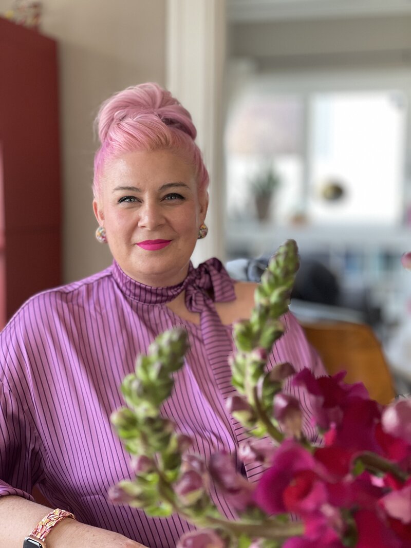 Woman with pink hair in a bun wearing a bright pink shirt sitting in a chair next to pink flowers.