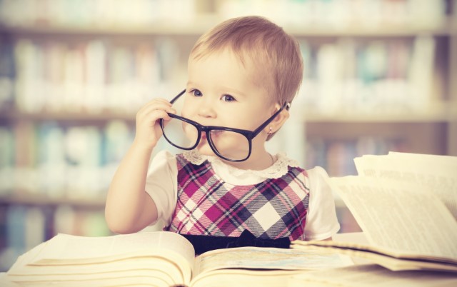 Toddler pulls off black rimmed glasses while sitting behind a big open book.