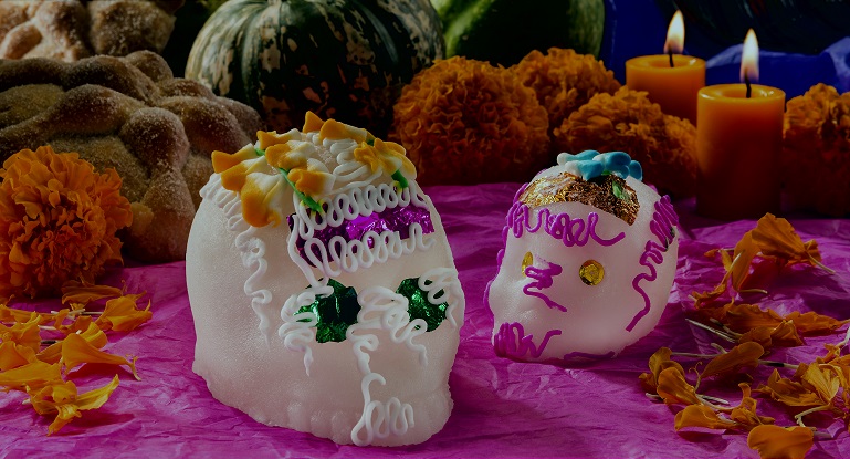 Image of white sugar skulls decorated with mulitcolor icing on a magenta table with orange candles and marigolds
