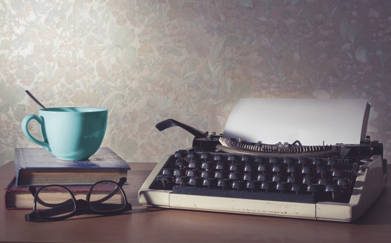 Image of typewriter next to a book with a blue cup on top 