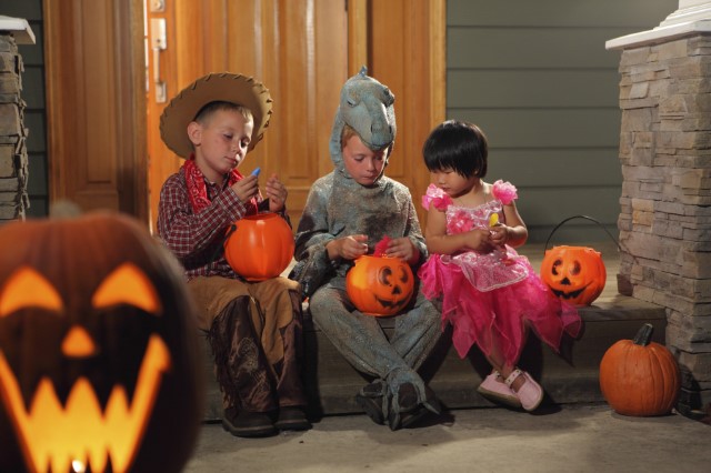 Halloween scene with three children in costume sitting on a porch holding their jack-o-lantern candy buckets