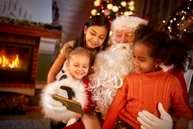 Santa Claus has a white beard, red/white hat, red suit and black belt, and white gloves. Santa is reading to children behind a green Christmas tree. 
