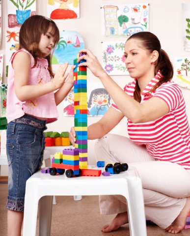 Parent in red/white striped shirt and white pants and child in jeans and pink shirt building with colorful LEGO blocks on a white table. 