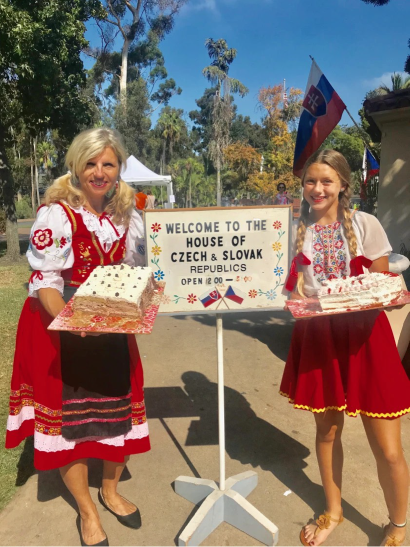 two women dressed in traditional costume holding cakes next to sign for House of Czech & Slovak Republics
