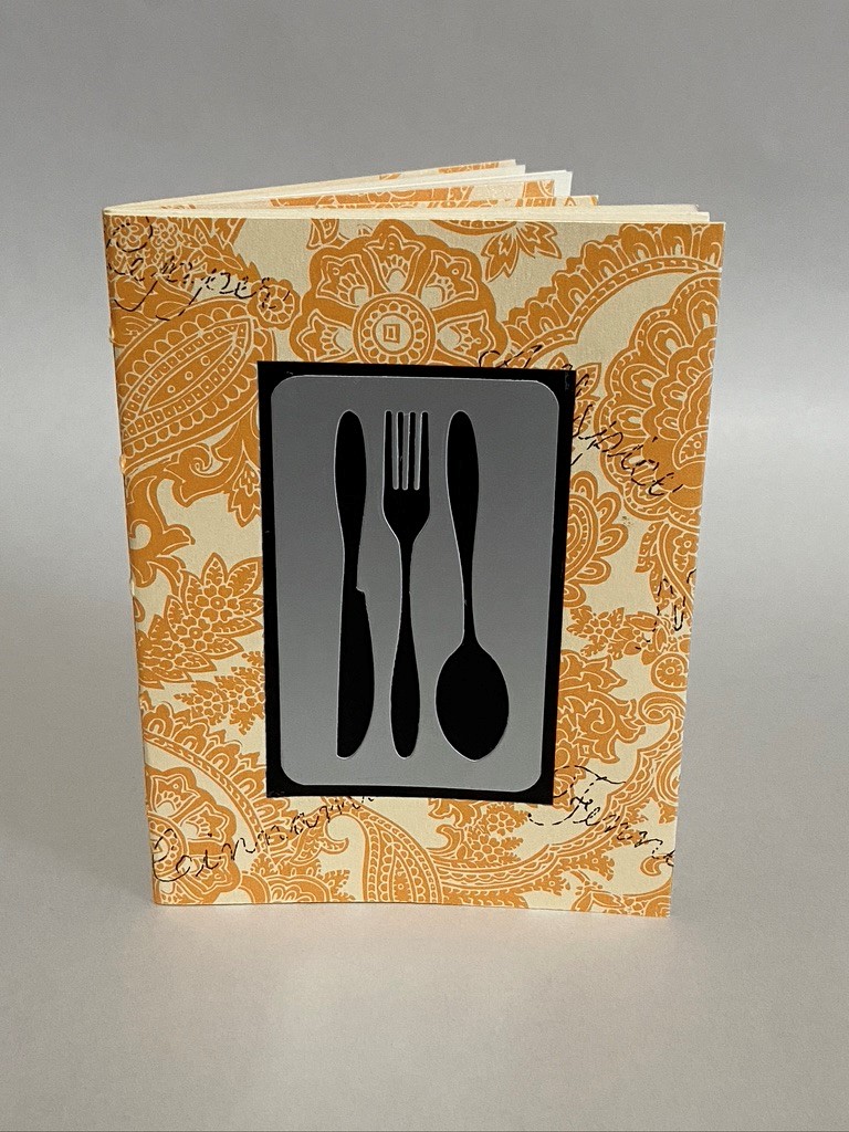 Hand made recipe book; on the cover is a decorative border with a center graphic of a knife, fork, and spoon