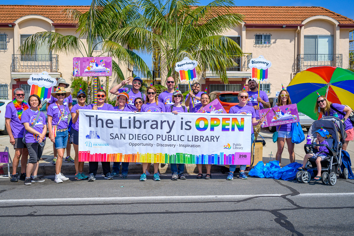 Photo of library staff and volunteers in purple pride shirts holding up banner that reads "The Library is Open."