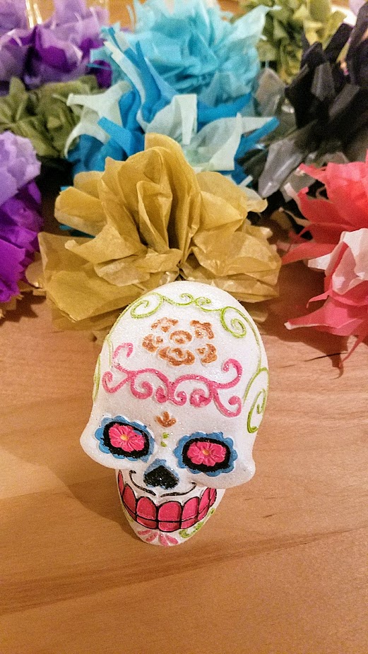 Picture of a white sugar skull with two large black eyes and a black smile, decorated with pink and orange icing squilly lines. The skull is on a brown table and yellow, pink, purple, and blue flowers are in the background. 