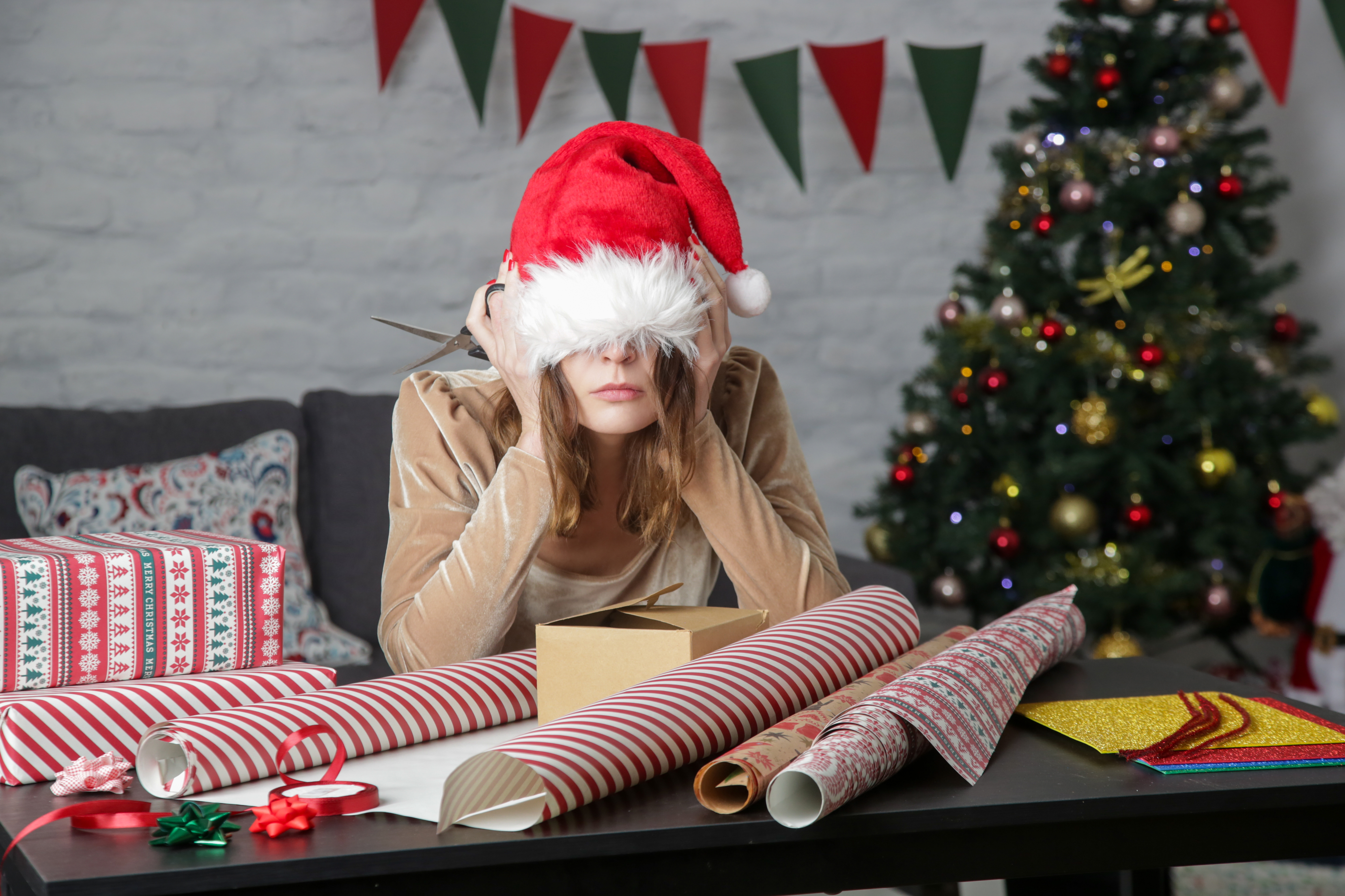 Holiday scene with decorated tree in background and woman with a Santa hat pulled over her eyes with piles of gift wrap