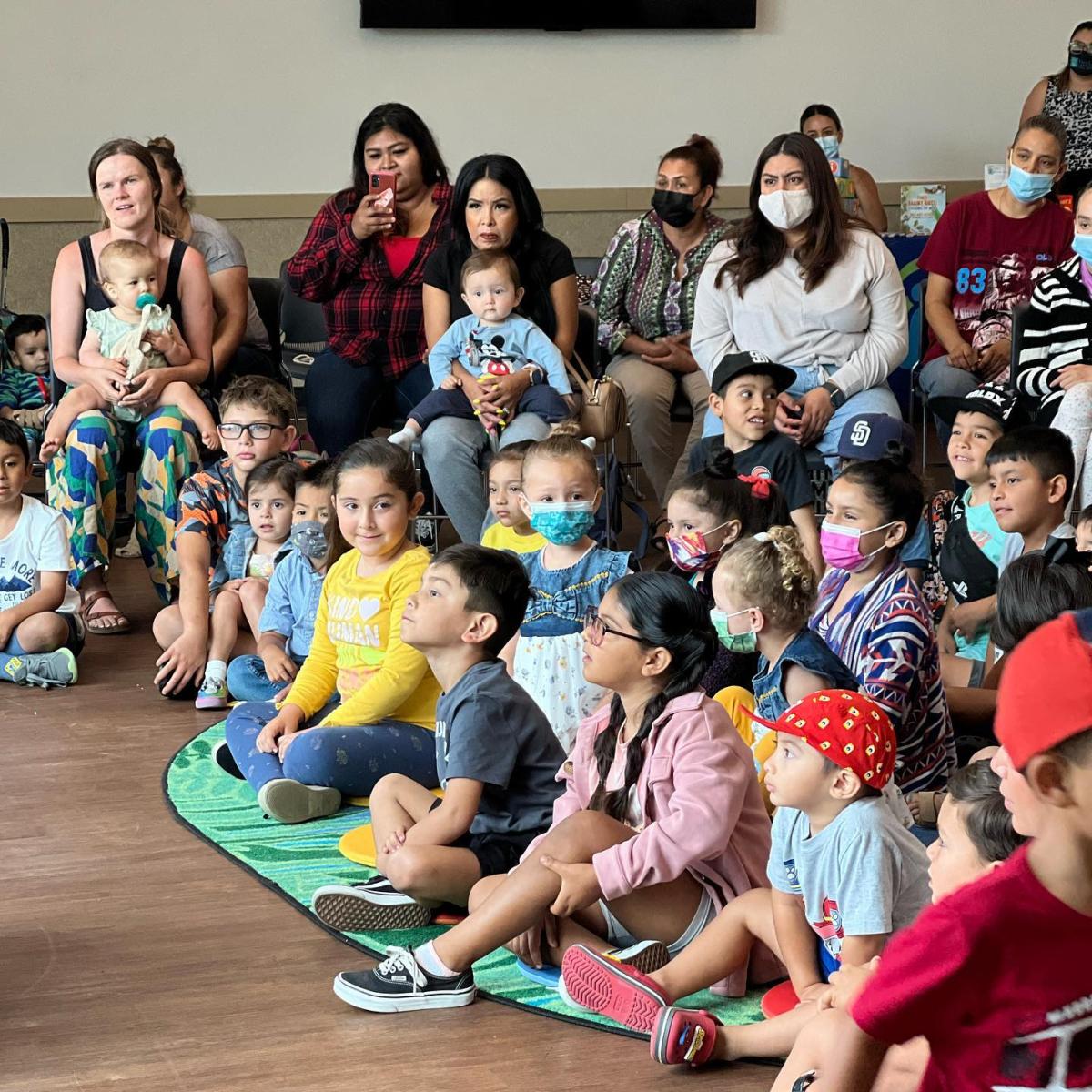 Image of people, kids and adults, with masks sitting on the floor listening