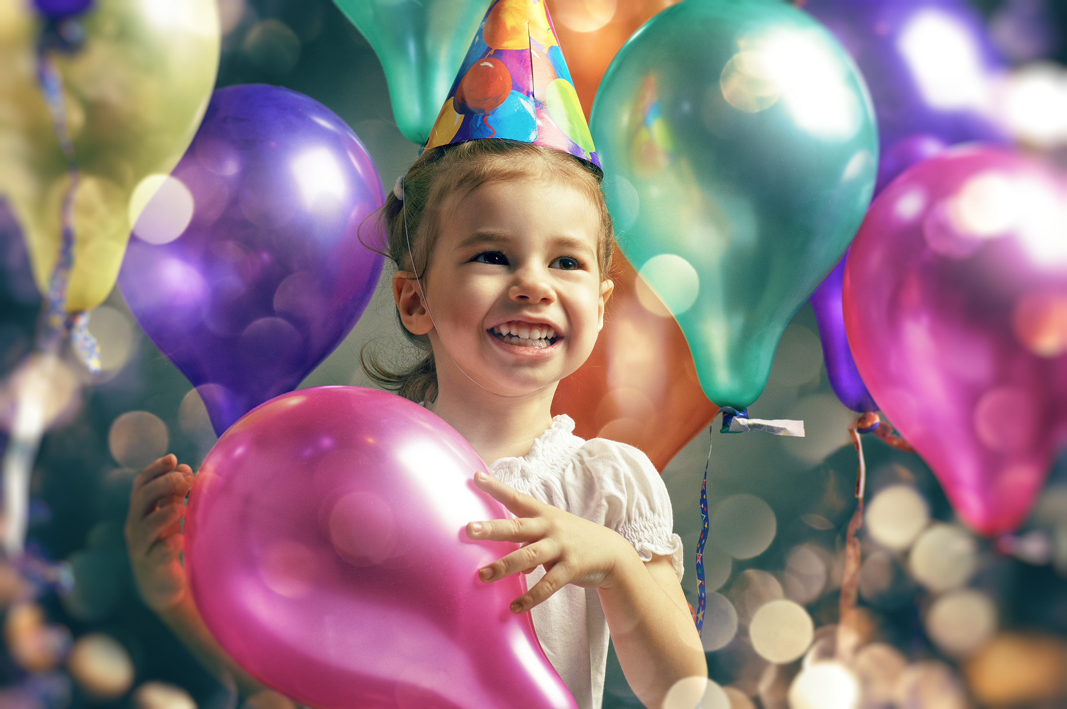 Young girl in party hat holding a pink balloon with multicolored balloons in the background