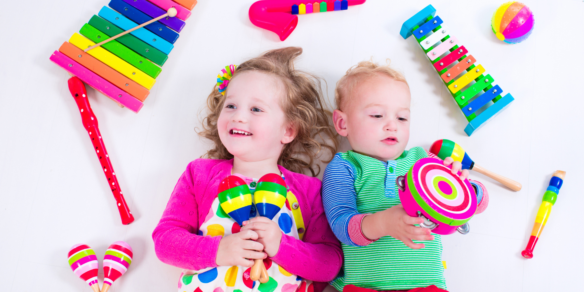 Photograph of 2 light-skinned toddlers lying on a white background surrounded by colorful musical instruments