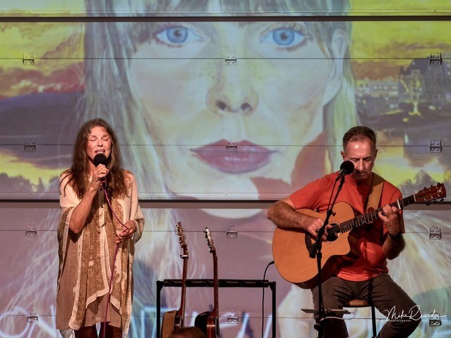 Photo of a female singer and man strumming a guitar, in front of a drawing of Joni Mitchell