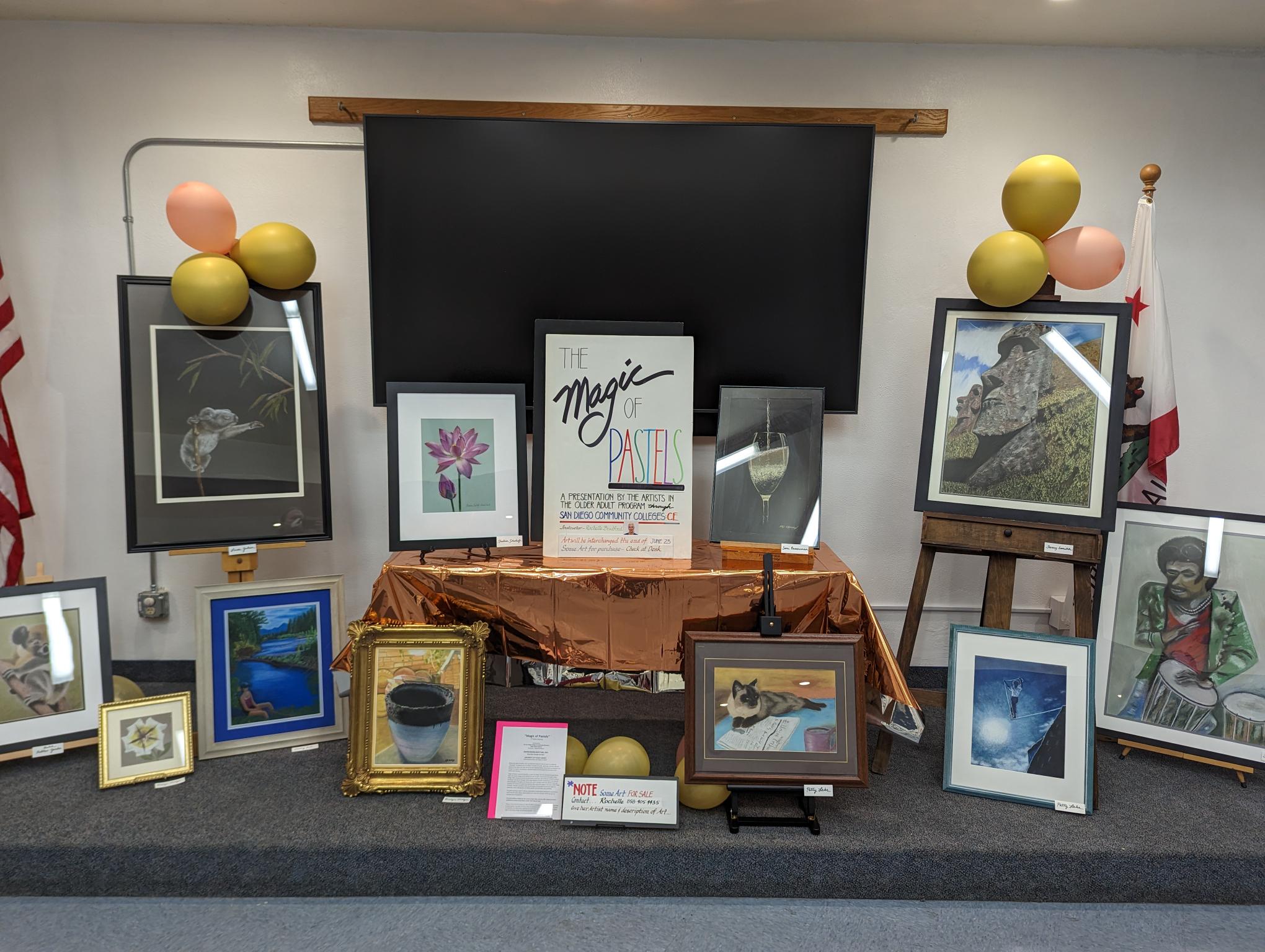 A display of pastel works including portraits, landscapes, and still lifes surrounding a sign with the title of the exhibition.  The sign reads "The Magic of Pastels. A presentation by the artists in the older adult program through San Diego Community College Continuing Education. Instructor Rochelle Bradford.
