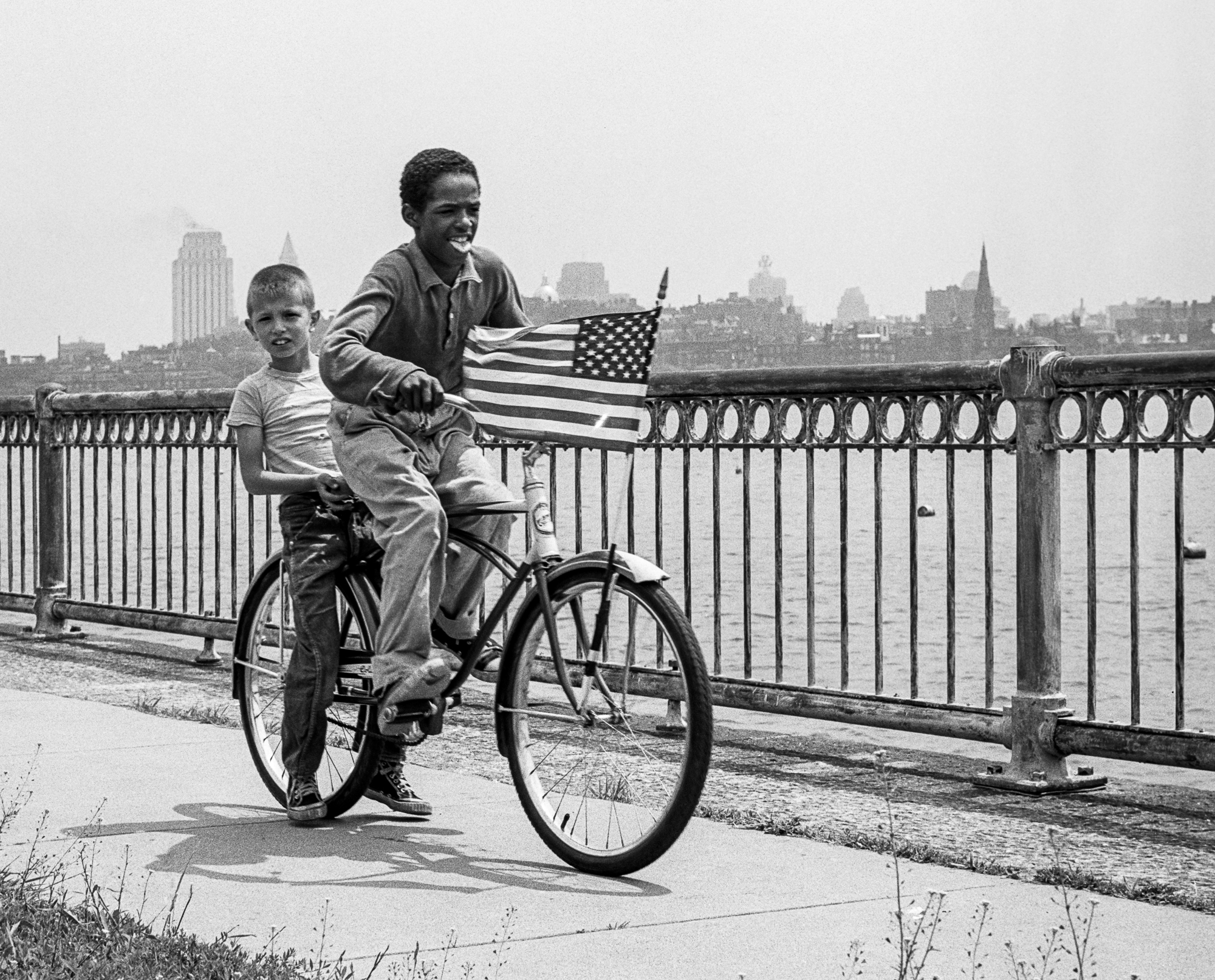 Black and white photo of two boys riding a bicycle in an urban landscape