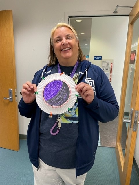 Ms. Maureen is holding an example of a tambourine made with paper plates and bells, decorated in purple