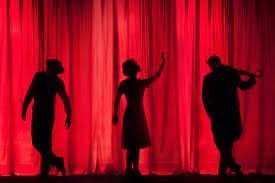 Three performers stand on stage silhouetted in front of a red curtain.
