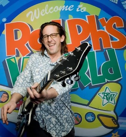 Man with long hair and glasses holding guitar and smiling