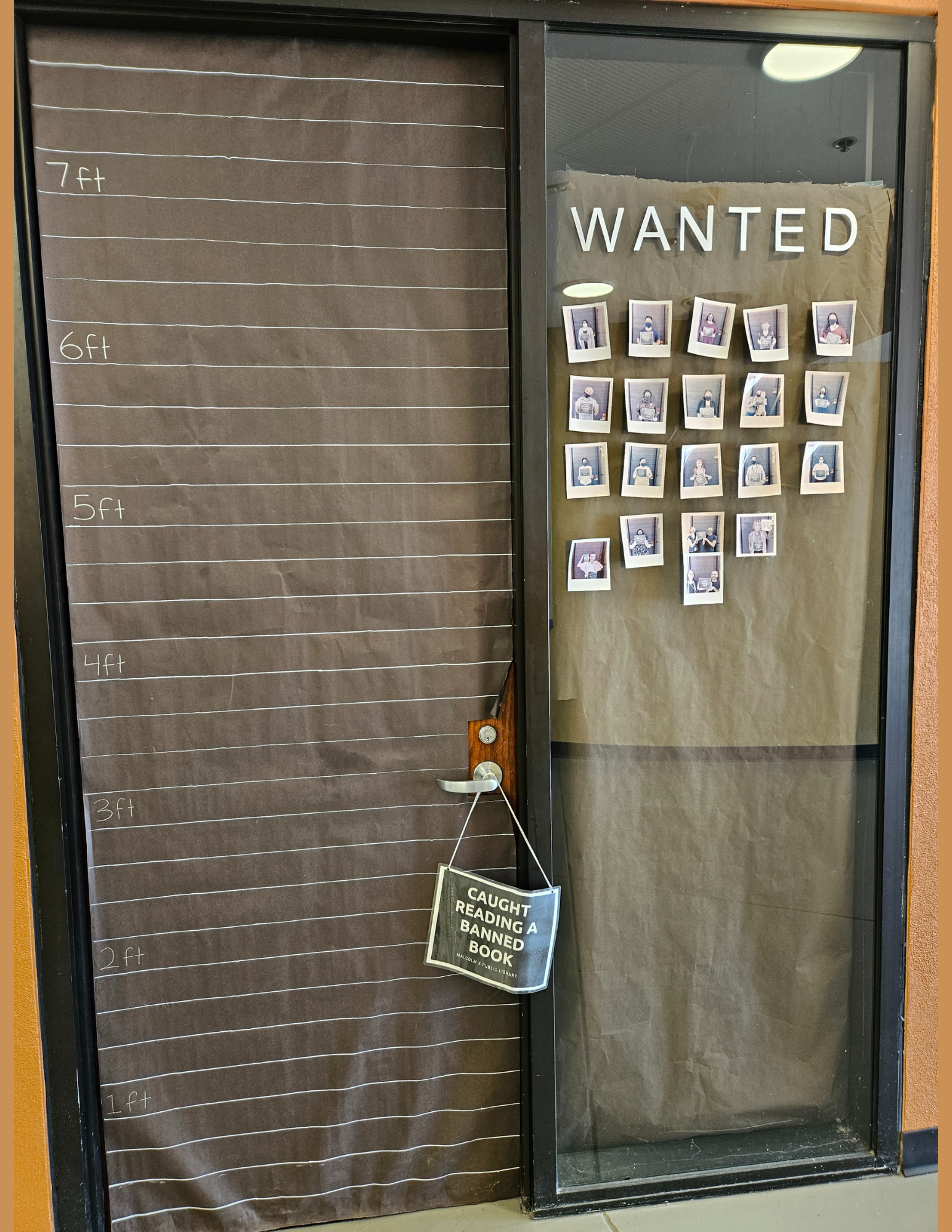 A photograph of the Banned Books Week Wanted Wall. A door is covered in black paper with white lines with numbers on the left side. A sign stating "caught reading a banned book" is hanging from the doorknob. The window to the right of the door says "wanted" and displays photos of people in front of the door.