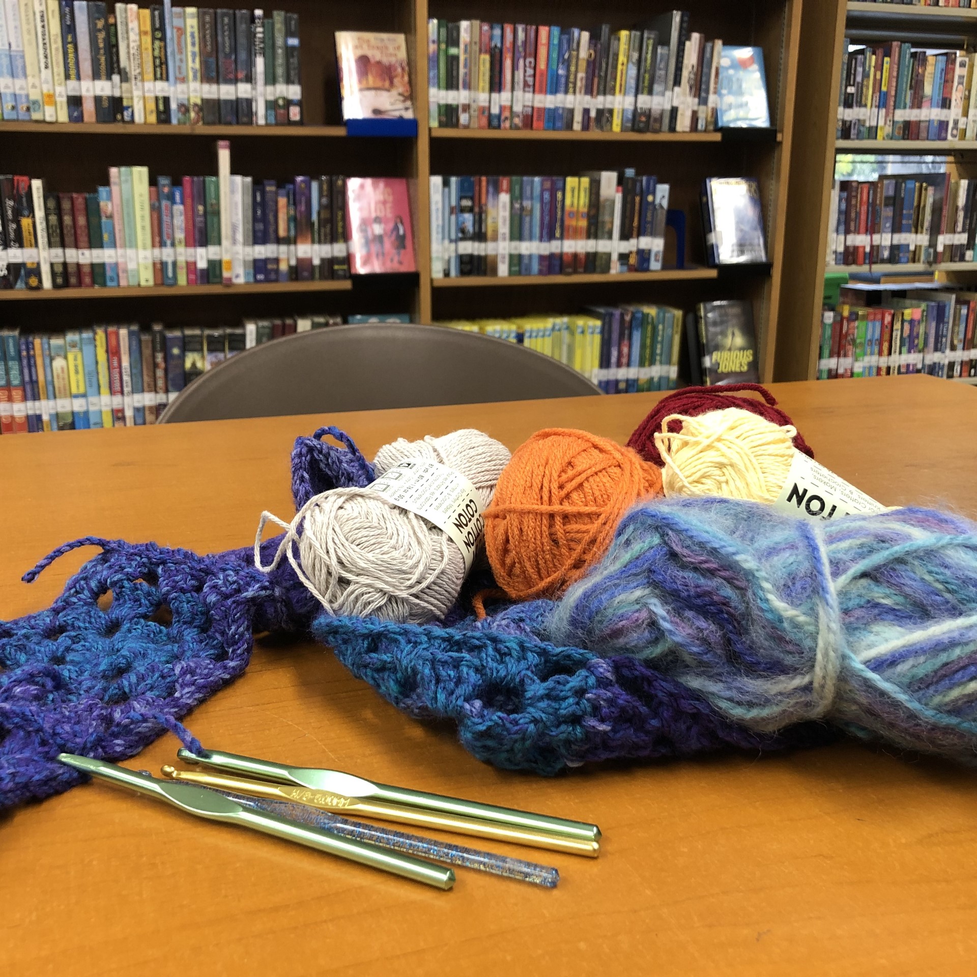 a pile of colorful yarn and some chrochet hooks on a table with a bookshelf behind it at the Kensington-Normal Heights Branch Library.