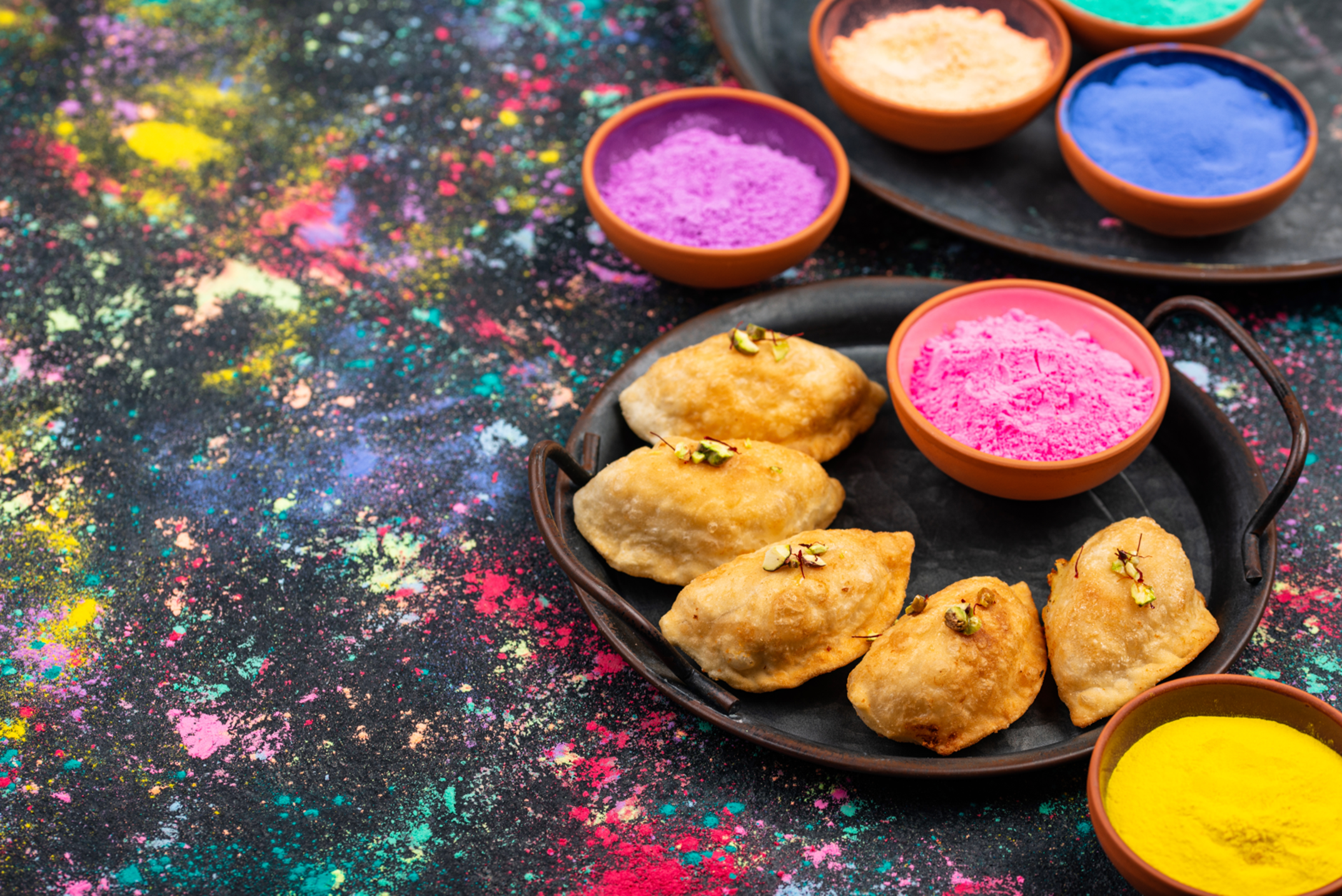 A picture of Gujiya on a plate with red, yellow, blue, and purple bowls of colored powder.