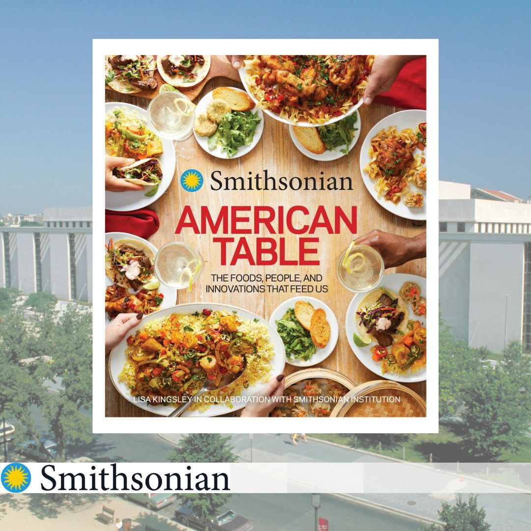 A book titled Smithsonian American Table, The Foods, People, and Innovations that Feed Us with images of foods including pasta, roast chicken, and tacos on it.