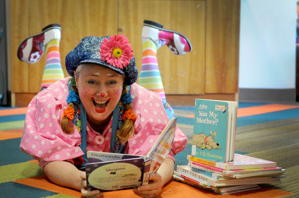 Clown in pink blouse and blue denim hat, reading a book on a carpet.
