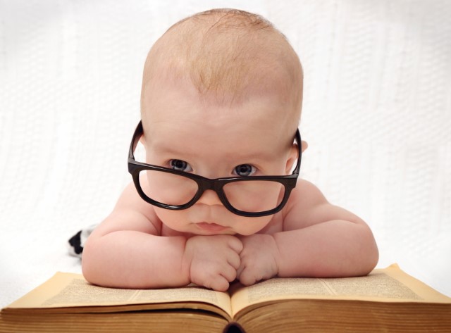 Bald baby wearing black framed glasses leaning on an open book