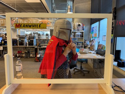 White frame that mimics a comic book panel with words "Meanwhile" in corner and woman wearing a 3D printed Mandalorian helmet posing