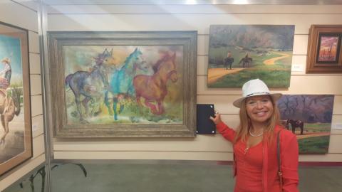 Woman wearing red top and white hat stands next to paintings, including a painting of horses galloping.