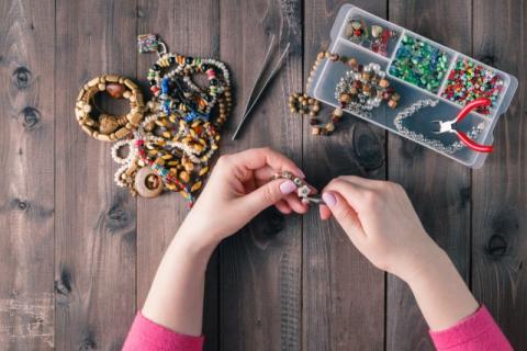 Image of person's hands as they are creating something with beads. Beaded jewelry is on the left of the screen while a plastic container containing beads and beadwork tools are on the right.