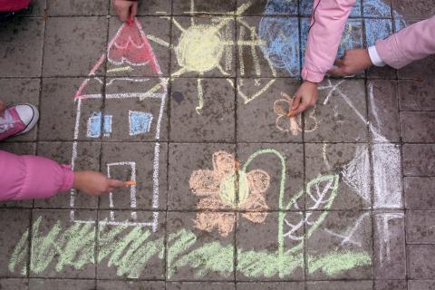 The arms of two children drawing a house, flowers and the sun with sidewalk chalk