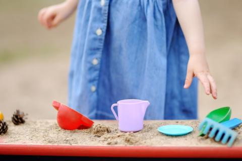 Child in blue dress stands behind a table with sand and cups and plates.