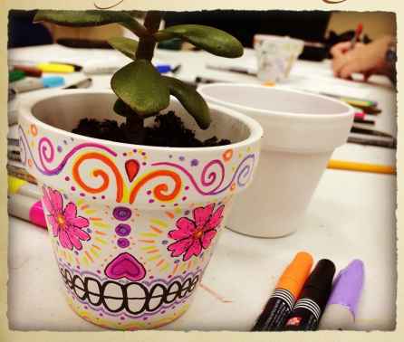 A white flower pot with a green succulent in it. A student is creating a skeleton face with pink flowers and purple teeth and other decorative designs in honor of Day of the Dead Latinx culture. 