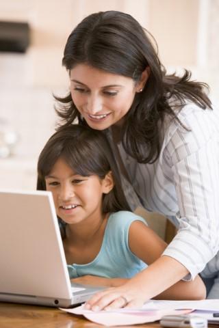 A middle-aged hispanic woman stands behind a young girl sitting in front of a computer