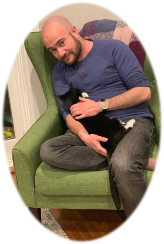 Image of Author Kyle Lukoff, author is wearing black jeans, a blue sweatshirt and is sitting on a neon green chair and holding a black cat in his lap.