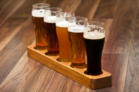 A "flight" of 5 small glasses of beer of different shades sit on a thin wooden serving tray.