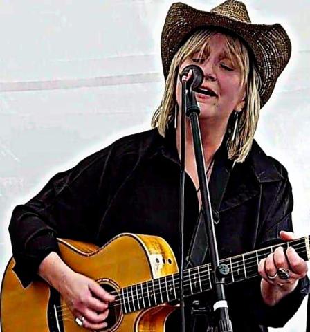Photo of a light-skinned woman with blond hair wearing a dark-colored shirt and cowboy hat. She is singing into a microphone and playing a guitar