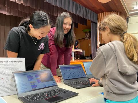 Two high school girls smiling at a younger girl who is working on a laptop.