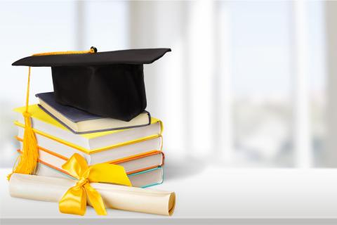 Black graduation cap on top of a pile of books, next to a roll of paper tied with a yellow ribbon, and on top of a white table.