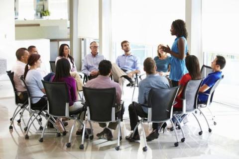 A circle of people sitting in chairs, listening to a standing person. 