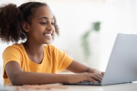 Girl about age 10 with brown skin and pigtails smiling and typing on laptop