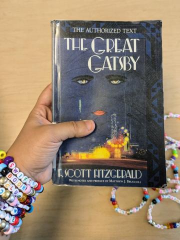 A hand with a wrist of friendship bracelets saying "Swiftie Book Club" holding this month's pick: The Great Gatsby.