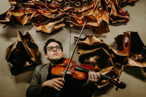 Photo of dark haired man with glasses holding a violin surrounded by wavy metal art