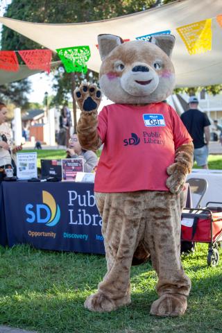 Odi The Coyote mascot has a red shirt on, he is standing against the library's outreach table with black table cloth on green grass. Colorful Papel Picado is hanging for this Day of the Dead celebration in Old Town San Diego.