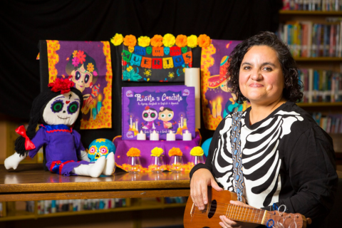 Ms. Jackie smiles while holding a ukelele. Colorful dia de los muertos accessories are displayed on a table behind her with the book Rosita and Conchita in the center.