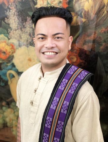 Professor Jojo's photo is on a fall colorful background. They are a traditional white Filipino long sleeve shirt and a purple scarf with intricate designs. 