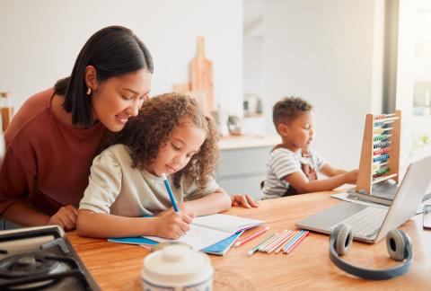 Mom with daughter, about age 6, in her lap working on homework. Son, about age 5, is playing with an educational toy at the table.