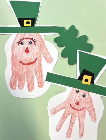 Paper crafts of colored in outlines of hands with faces drawn on. Green hats glued on.