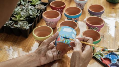 Hands painting a small terra cotta pot with pots and plants on the table in the background